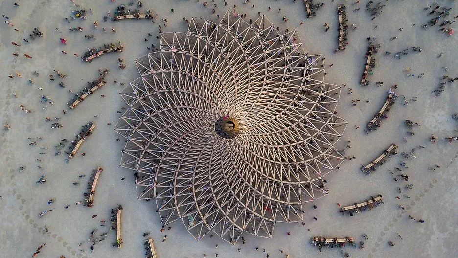 Aerial view of a large, spiral wooden structure with many people gathered around it on a light-colored ground.
