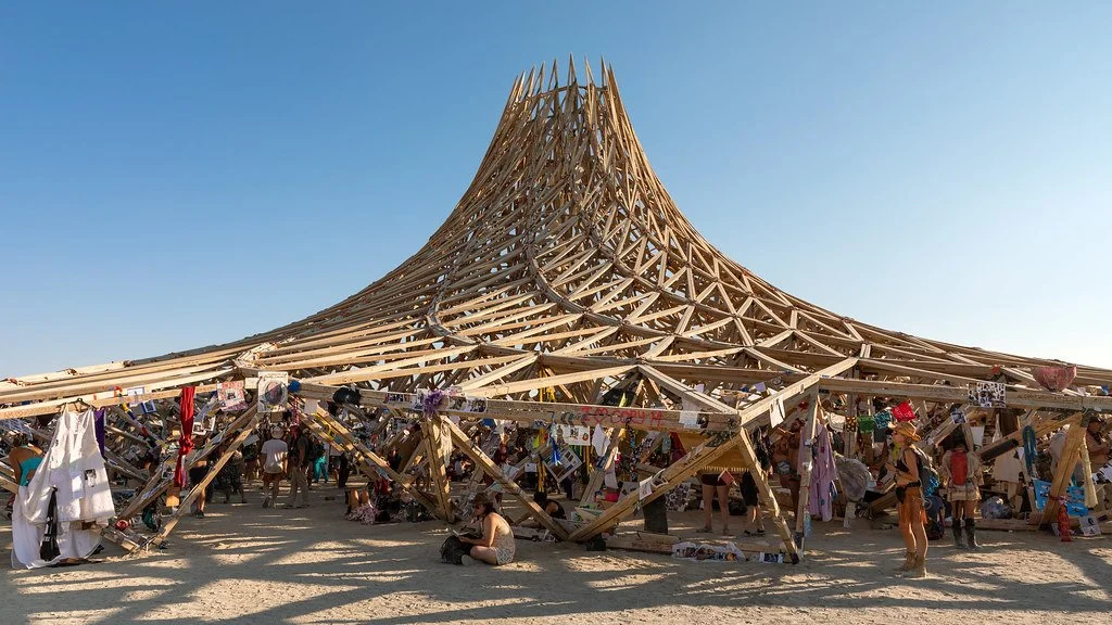 A large wooden conical structure under construction at the Burning Man festival, with many people around and colorful decorations.