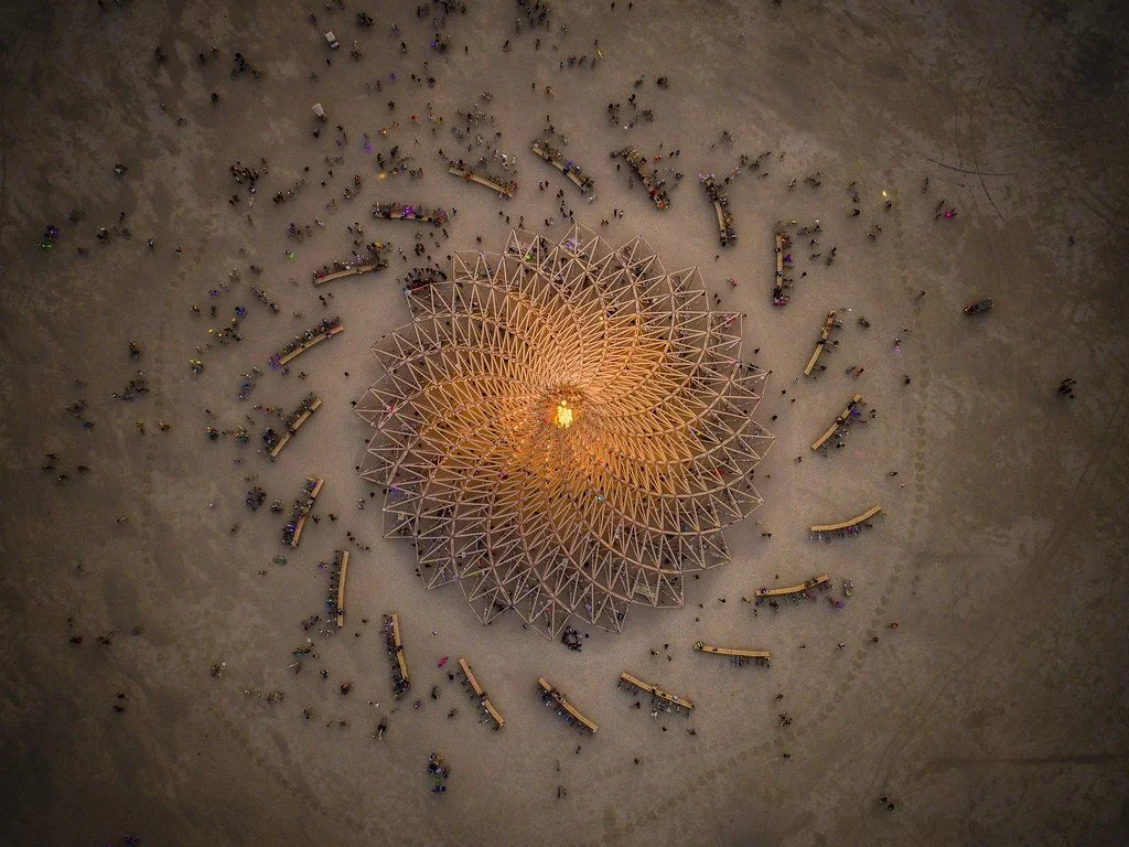 An aerial view of a large geometric wooden sculpture illuminated from the center, surrounded by numerous small people and wooden benches on a dirt ground at dusk or night.