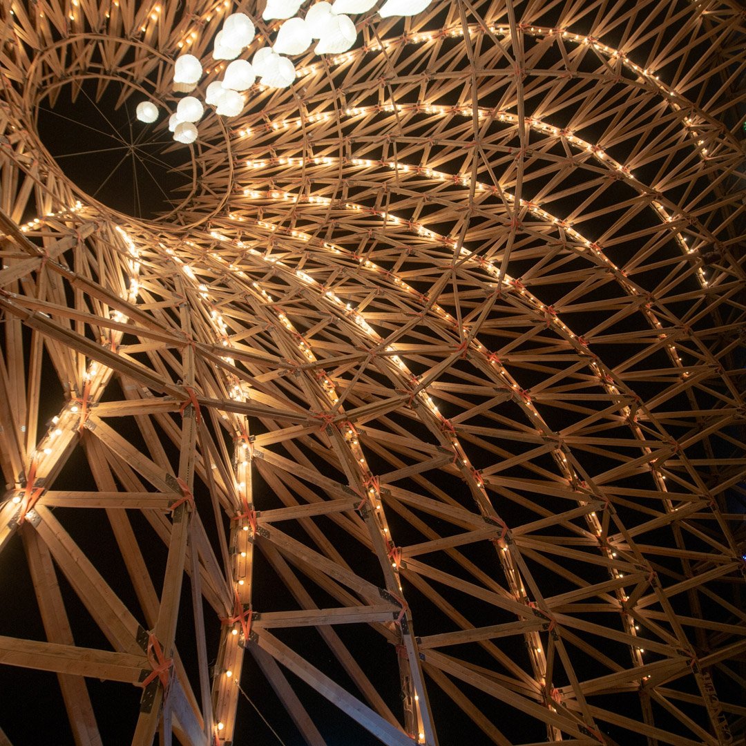 Close-up view of a wooden geometric sculpture illuminated with string lights at night.