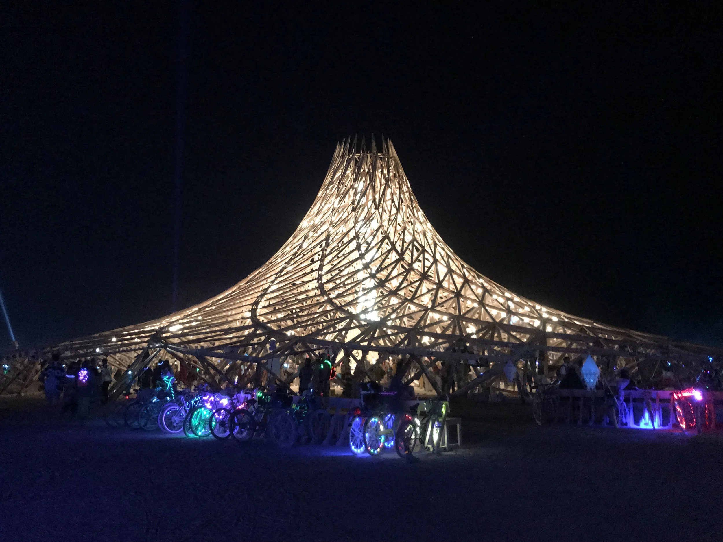 Night view of a large illuminated wooden lattice structure resembling a conical shape, with numerous people and colorful bicycles at its base, under a dark, starry sky.