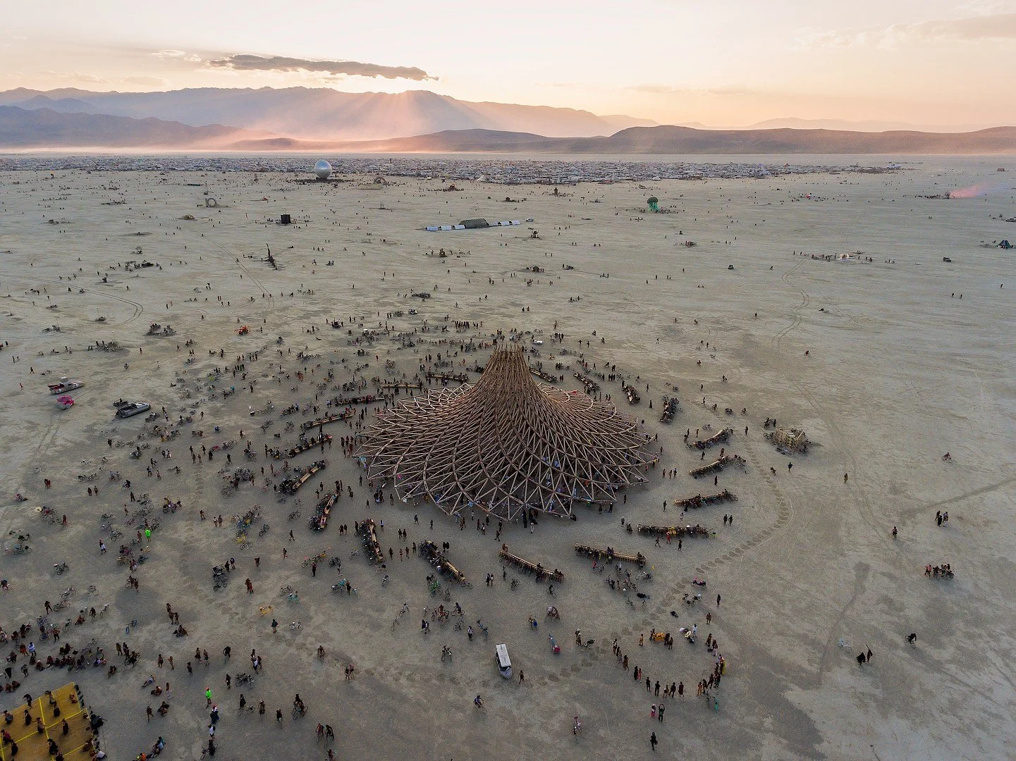 Aerial view of Burning Man festival with large wooden structure in desert, surrounded by people, vehicles, and artworks, with mountains in background.