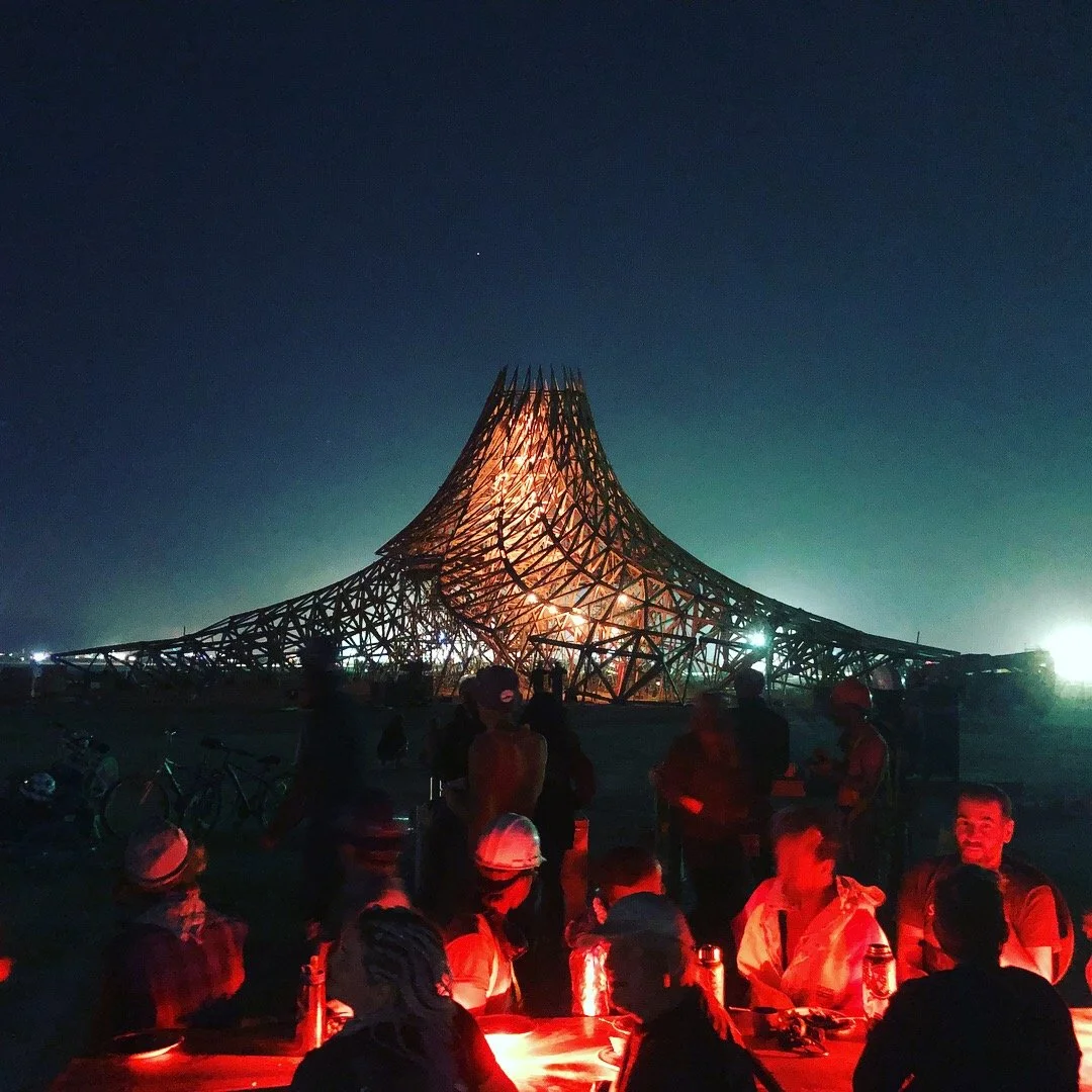 Night scene of a large wooden structure with a tent-like shape, illuminated from within, with people gathered at tables in the foreground.