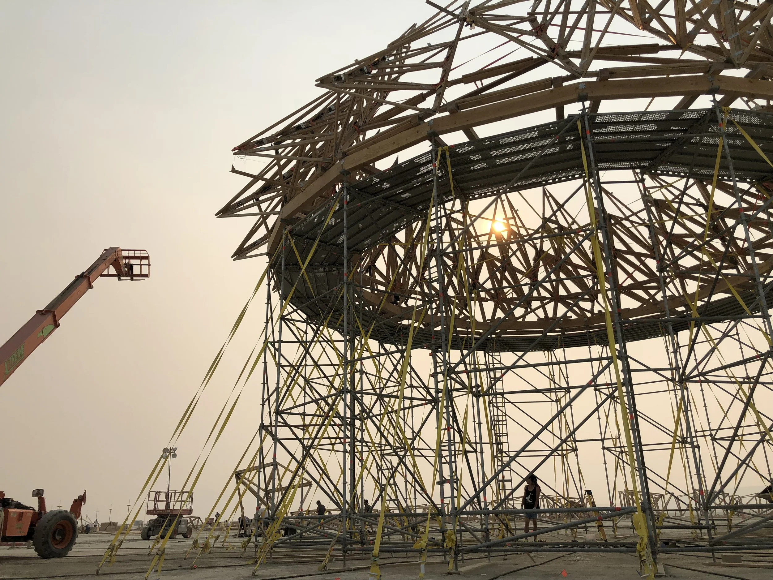 Construction workers building a large wooden structure, possibly a tower, with scaffolding and a crane nearby during sunset.