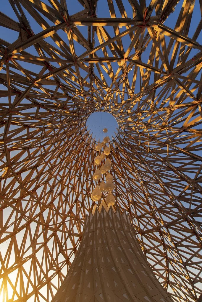 Looking up at a large geometric wooden tower with a circular opening at the top and floating lights hanging down, with the evening sky in the background.