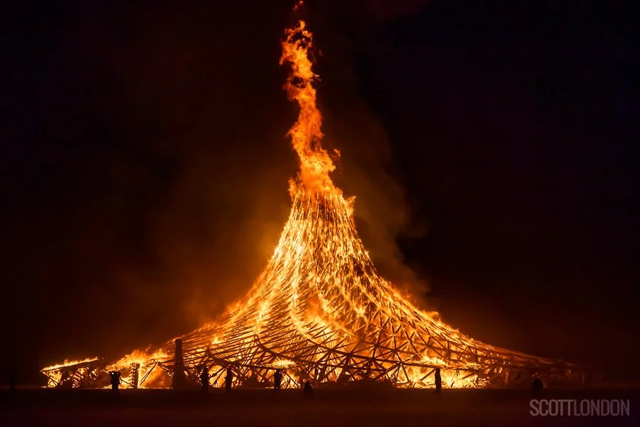 Large wooden structure on fire at night, with dark sky background and flames shooting upward.
