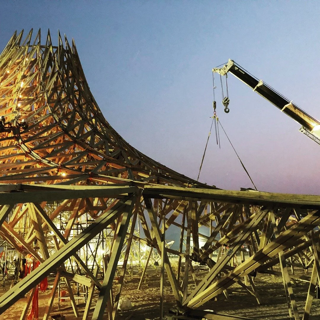 Construction site with a large wooden structure being built, illuminated with lights, and a crane lifting a section of the structure against a twilight sky.
