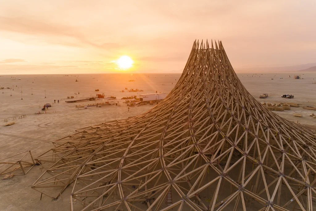 A large wooden structure under construction in a desert at sunset with the sun near the horizon. The structure has a tall, conical peak and a wide, spreading base, made of interlocking wooden beams. Scattered construction equipment and vehicles are v