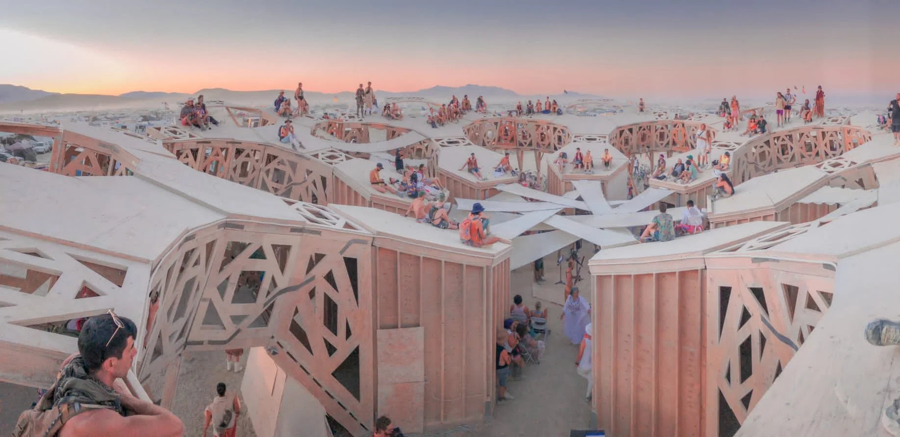 People sitting and walking on a modern wooden architectural structure with geometric patterns, during sunset with mountains in the background.