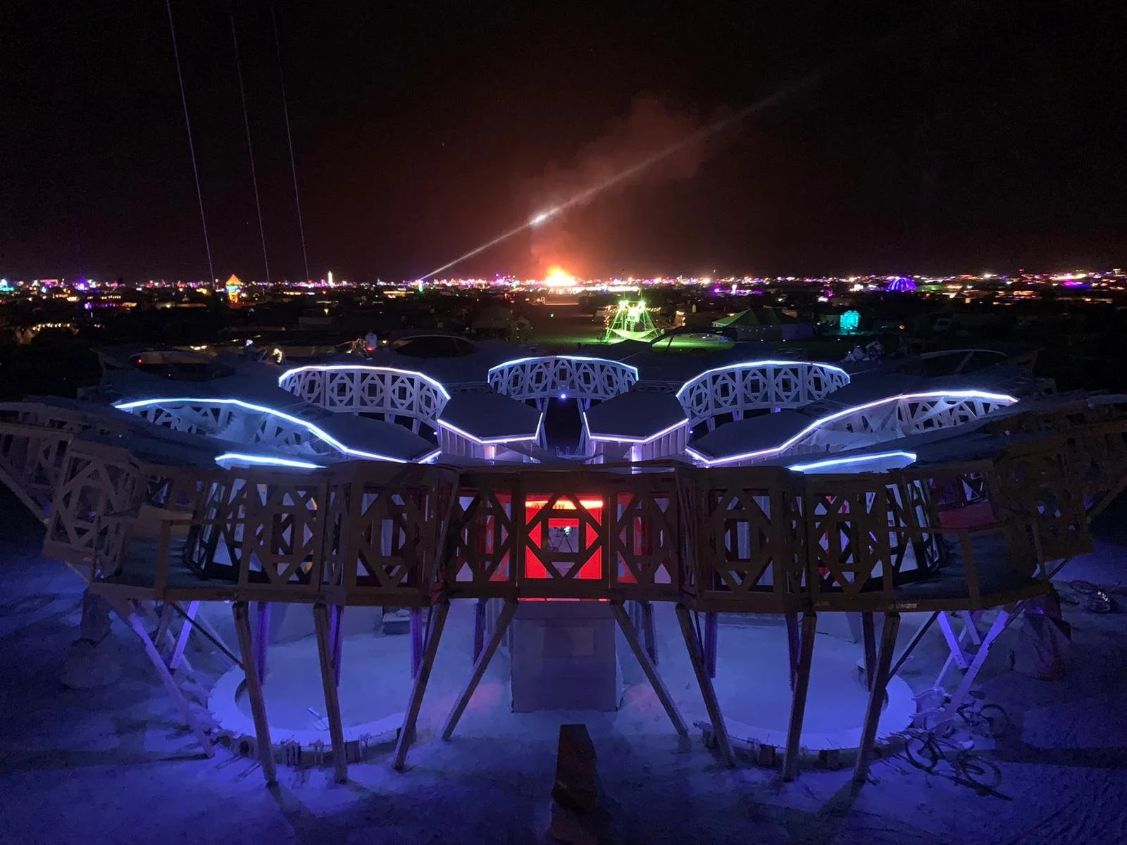 A night scene at Burning Man festival with a futuristic, neon-lit structure in the foreground. In the distance, an explosion and a bright streak of lightning can be seen against the dark sky, with colorful lights illuminating the landscape.