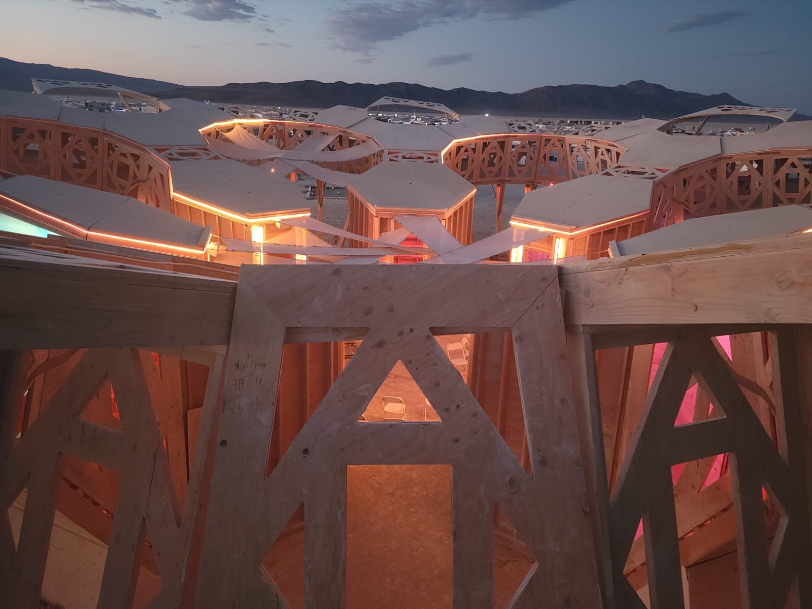 This image shows a wooden architectural structure under construction, with multiple interconnected round and angular segments outlined with lighting. The background features a landscape with mountains at dusk, and a partly cloudy sky.