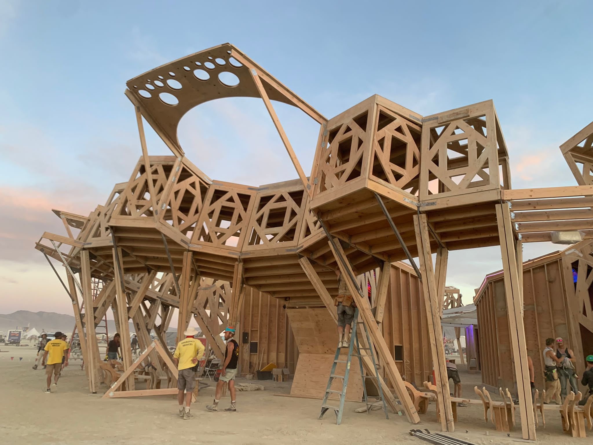 Wooden structure under construction with multiple people working and observing, possibly at a festival or event, with a clear sky and distant mountains in the background.