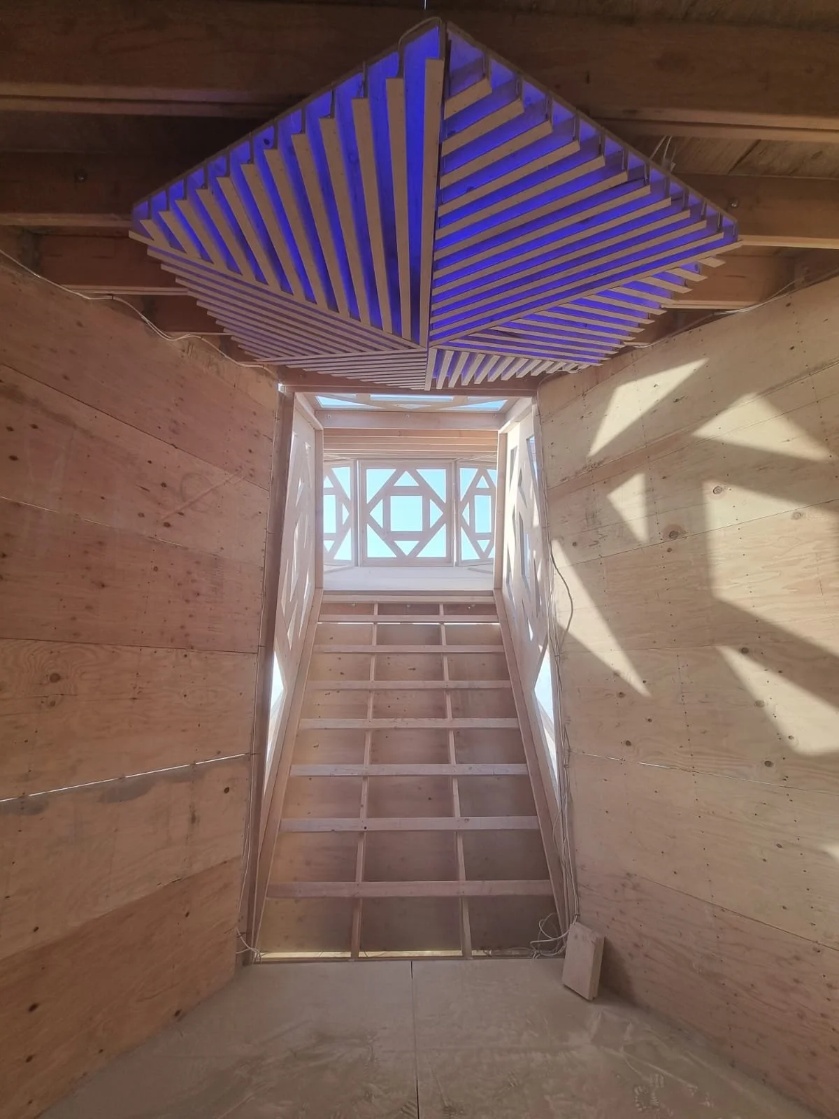 Interior view of a building under construction with wooden walls, a staircase opening, and a ceiling with wooden slats. There is a decorative window at the top and a blue-lit wooden structure hanging from the ceiling.