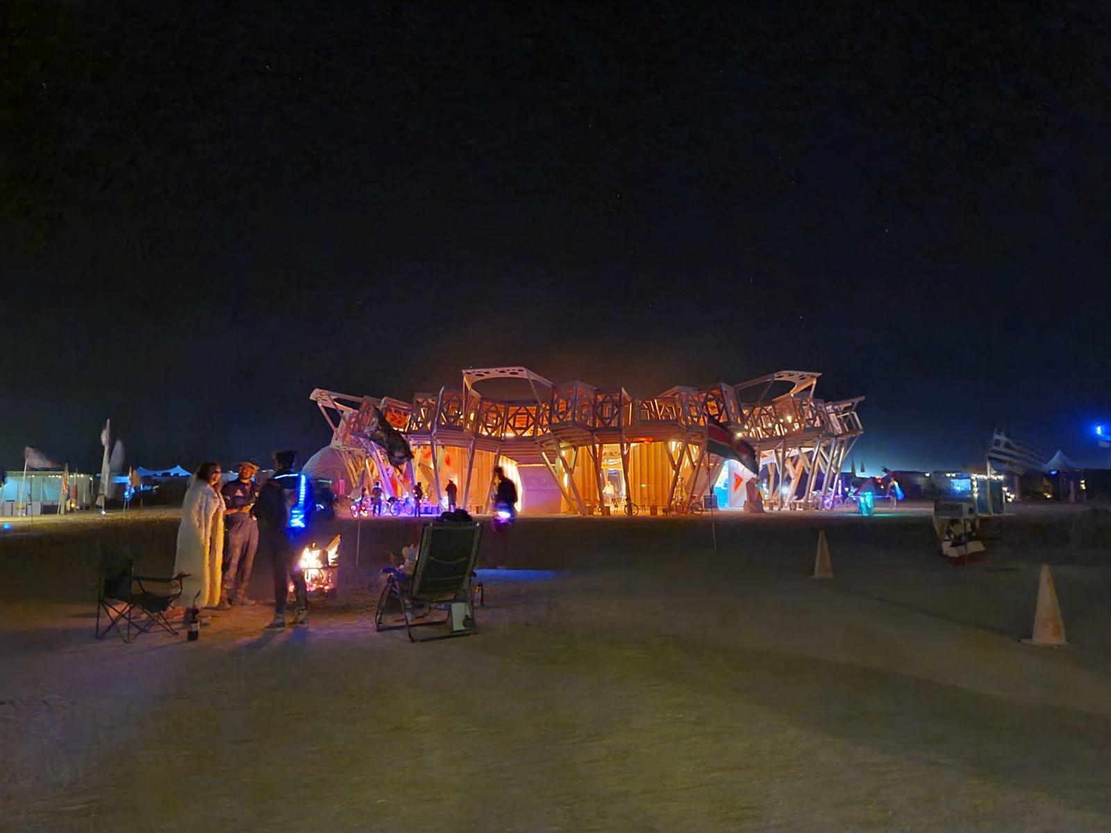 Nighttime scene at Burning Man with a large illuminated art installation resembling a ship, people gathered around sitting chairs, and tents in the background under a starry sky.