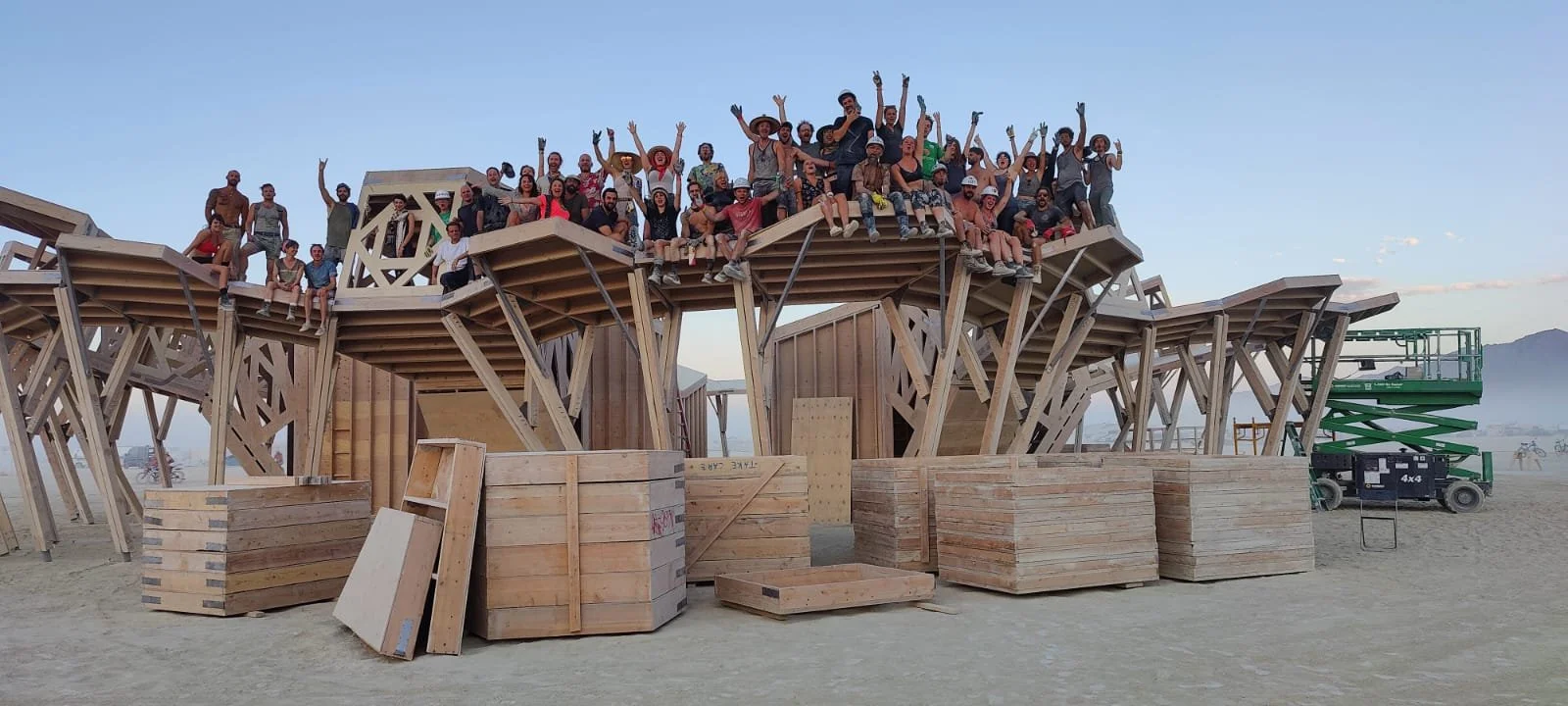 A large group of people sit and stand on a wooden structure resembling a futuristic, architectural design. The structure is under construction, with stacks of wood and construction equipment nearby, on a desert or dry lake bed.