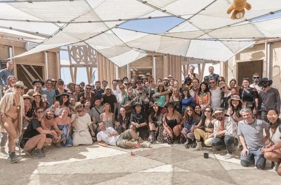 Large group of people posing for a photo on a construction site with wooden framework and partially built walls, under a makeshift canopy, in a desert-like environment.
