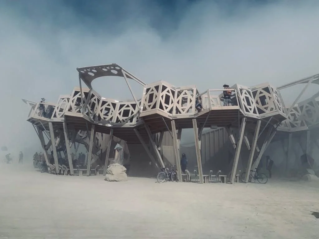 A modern, geometric observation tower with an elevated walkway in a desert environment, surrounded by dust and visitors.