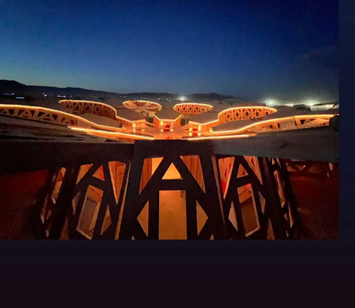 Illuminated sculpture structure at night in a desert landscape, with distant hills and a dark sky.