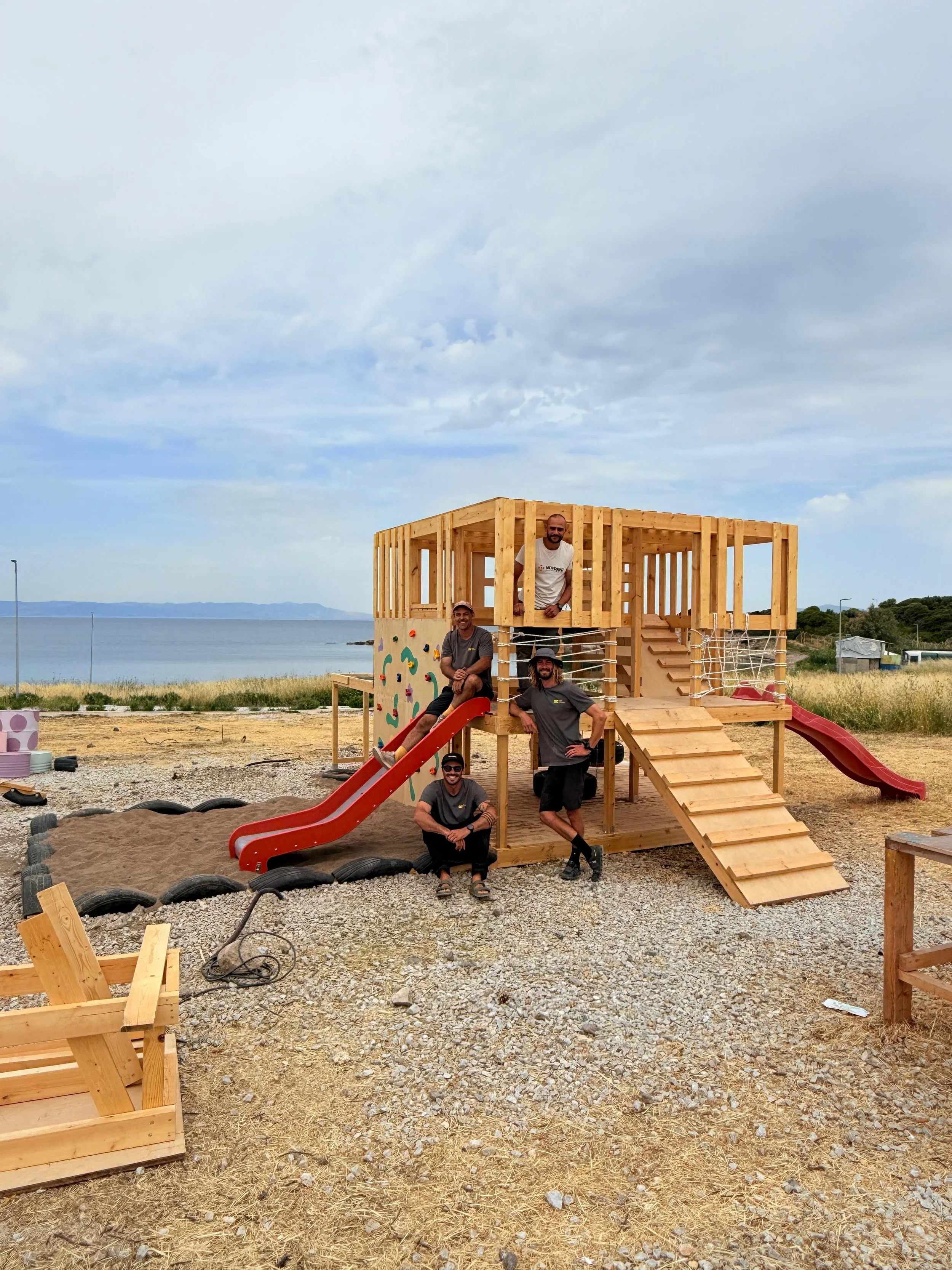 Four men building a wooden playground structure by the beach on a cloudy day.