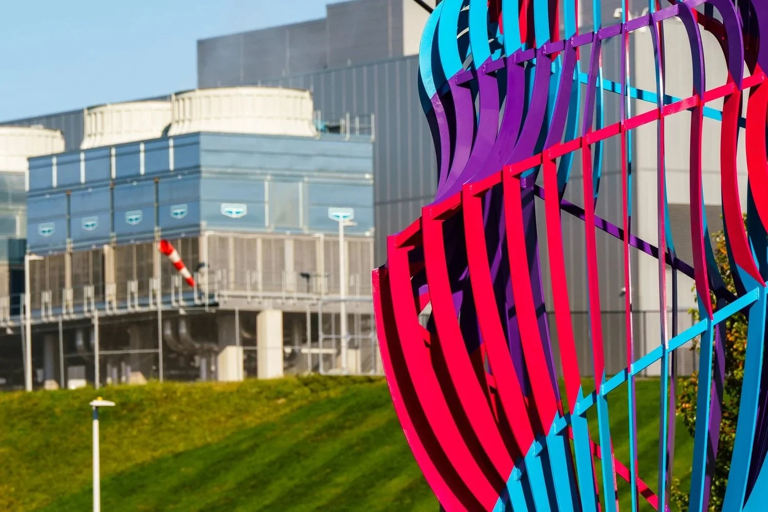 Colorful abstract metal sculpture in front of modern building with glass and metal facade, and grassy area with a streetlamp.