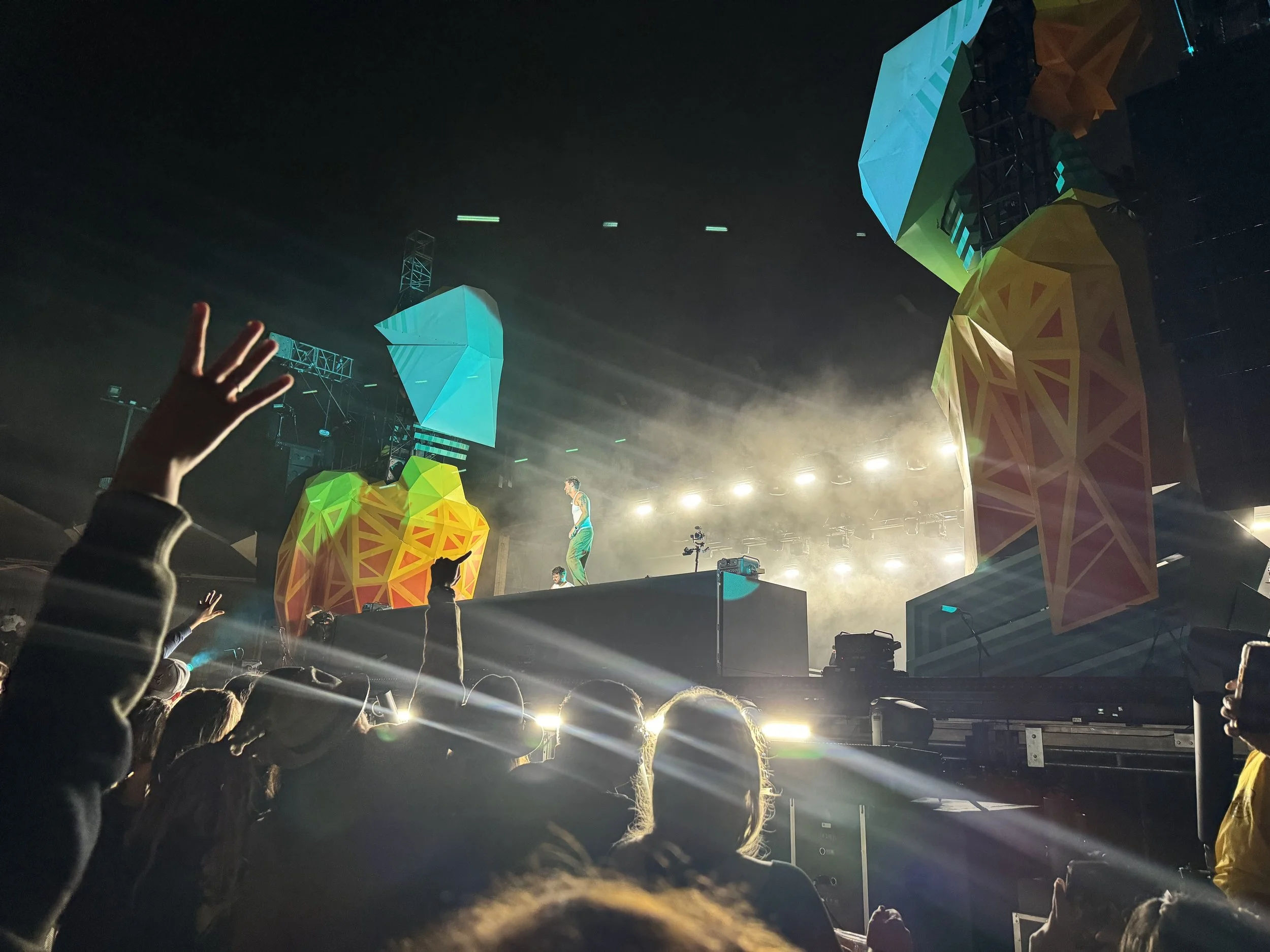 This image shows a concert or festival with a large crowd in front of a stage. The stage has colorful geometric decorations and large screens. There are performers on stage, and the setting is dark with bright lights illuminating the scene.