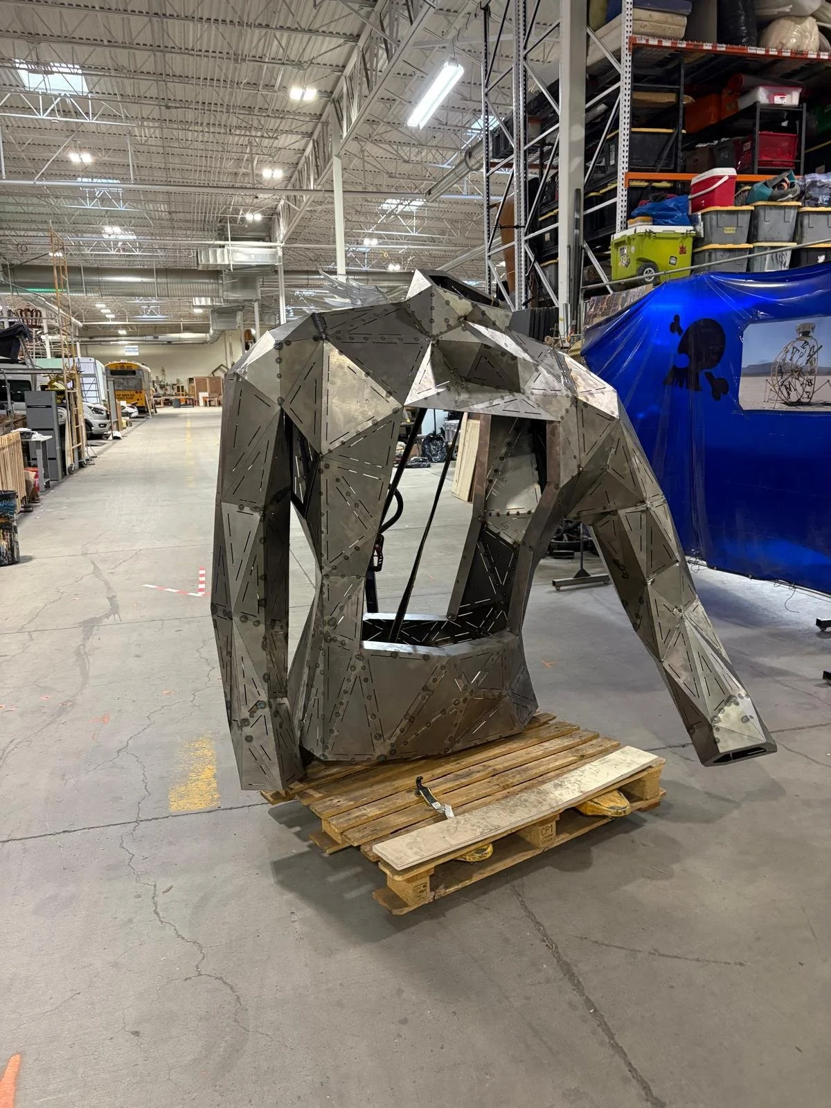 A large metal bear sculpture in progress on a wooden pallet inside a warehouse or workshop.