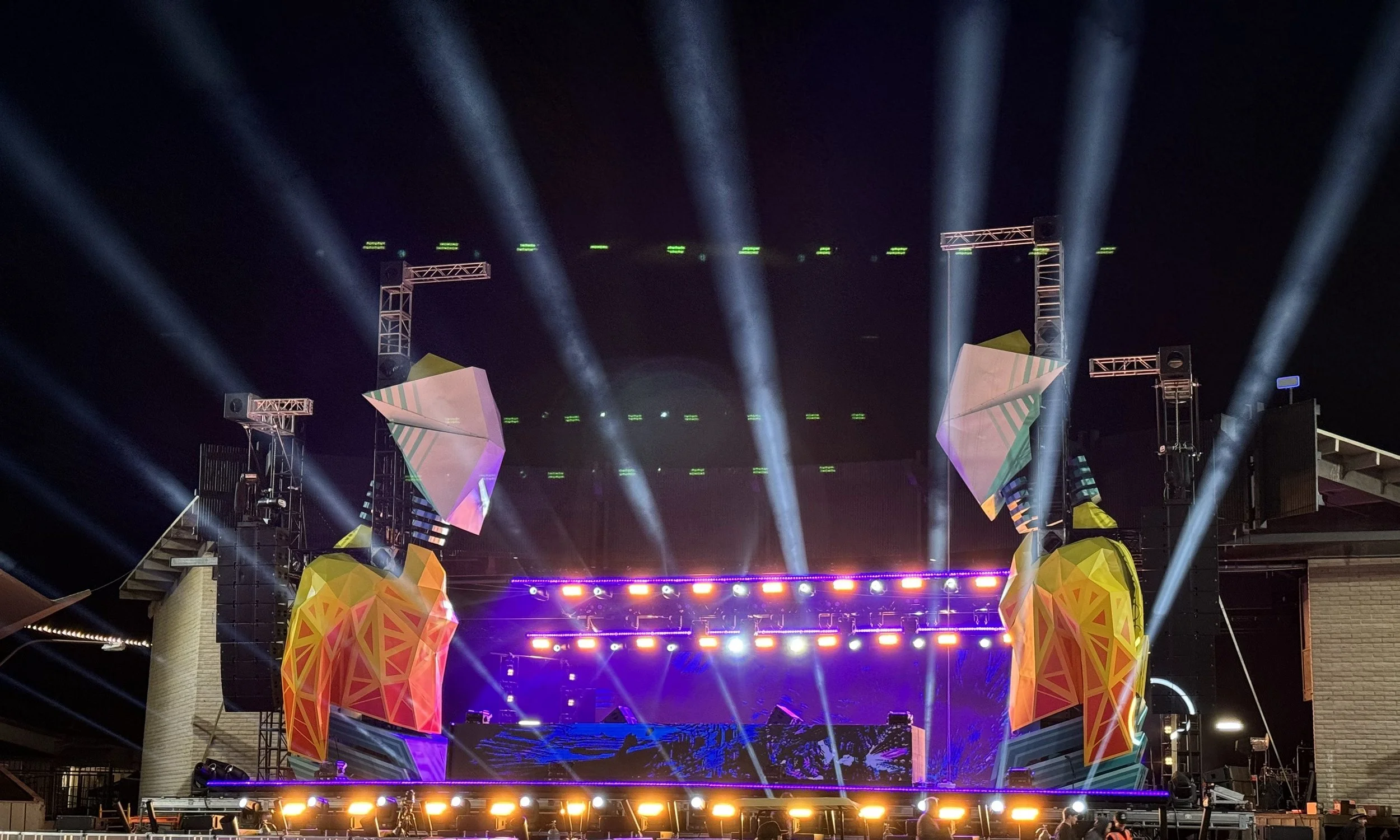 A vibrant outdoor concert stage at night with colorful geometric decorations, LED lights, and bright spotlights shining into the sky.