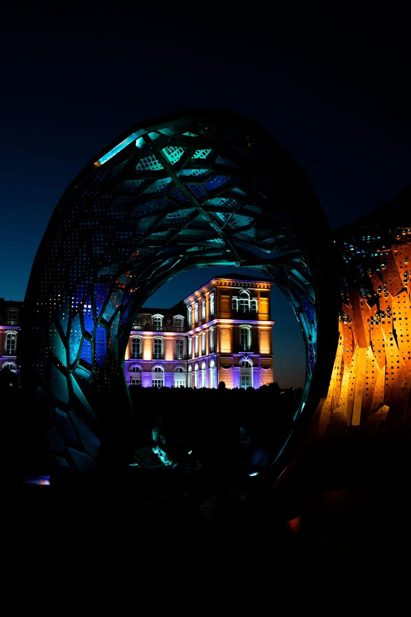 Night view of an illuminated architectural structure with a building in the background, featuring colorful lights and geometric design elements.