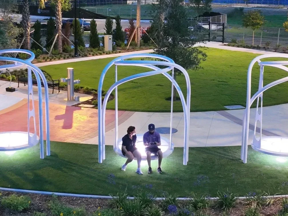 Two people sitting on modern swing seats in a park, illuminated at dusk, with greenery, pathways, and trees in the background.
