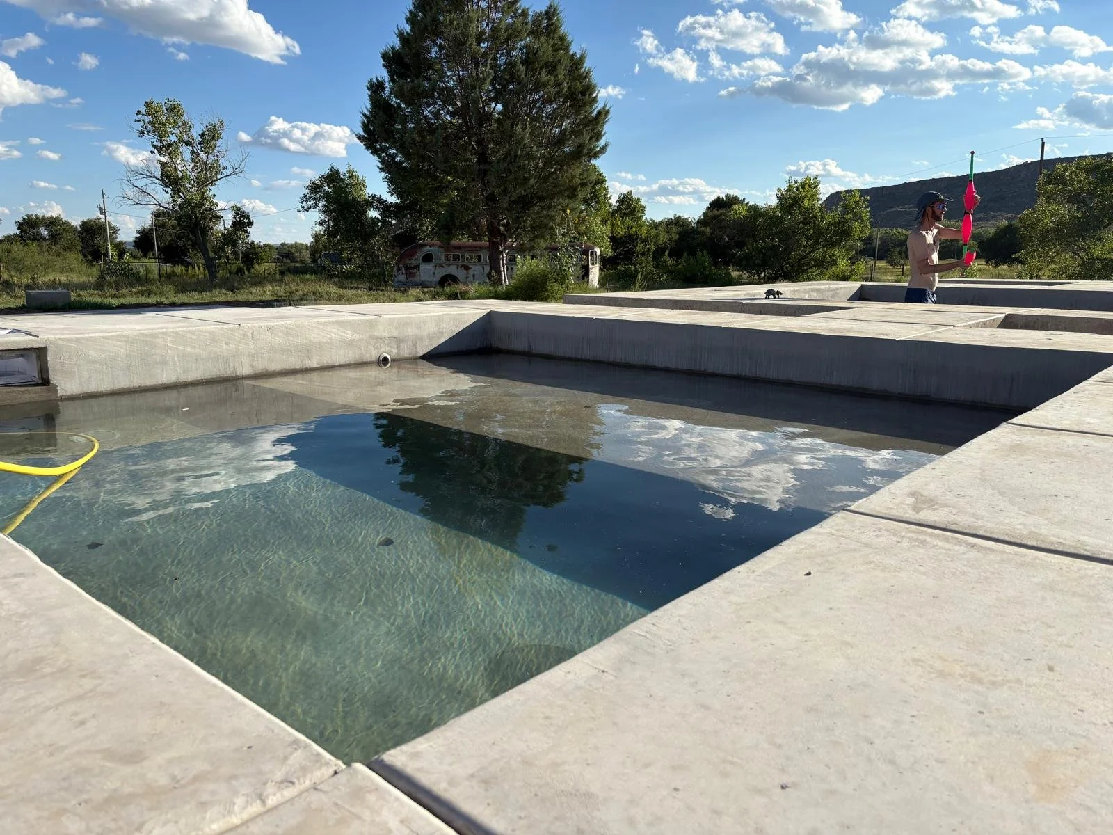 An empty concrete hot tub with water and a yellow hose inside, outdoors on a sunny day with trees, a vintage bus, and mountains in the background.