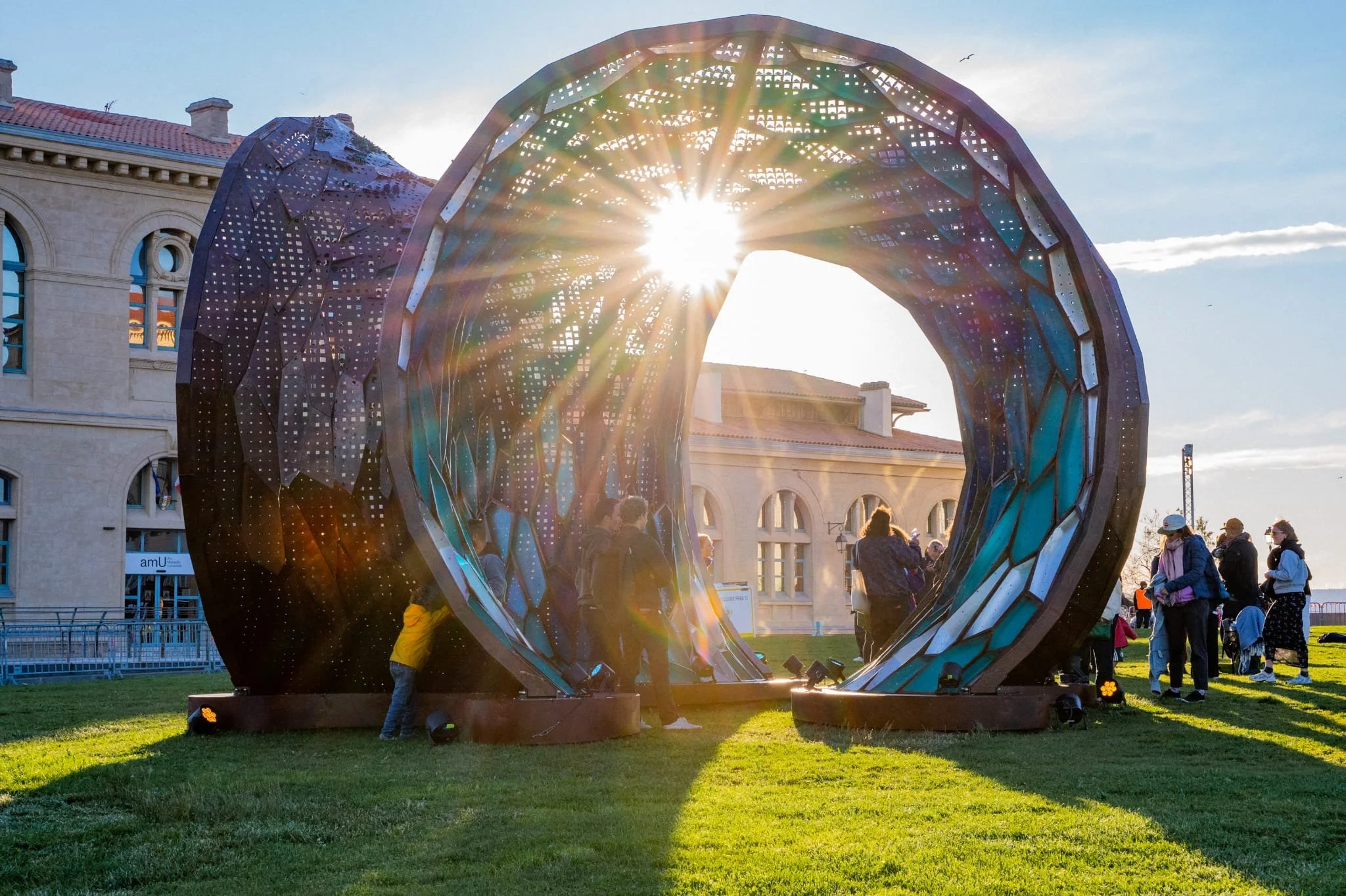 People gathered around a large, modern sculpture shaped like an open heart, with the sun shining through, creating a rainbow of light.