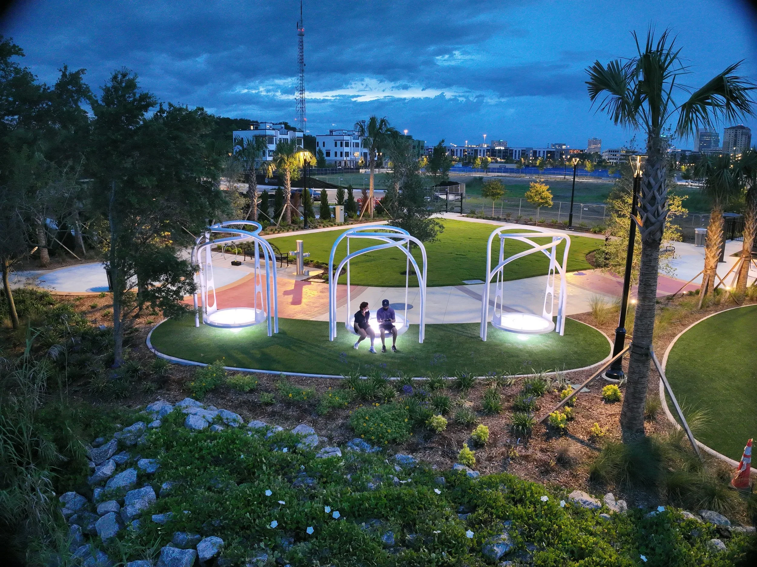 A park at dusk with modern white swings, palm trees, and two people sitting on a swing under lighting, surrounded by landscaped greenery, with a city skyline and cloudy sky in the background.