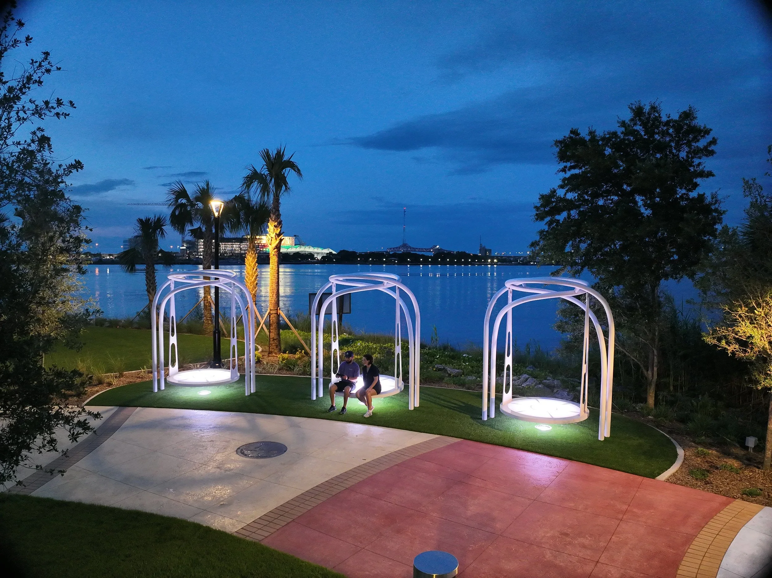 Two people sitting on a swing seat near a waterfront with palm trees, lit by lamps, under a blue evening sky.