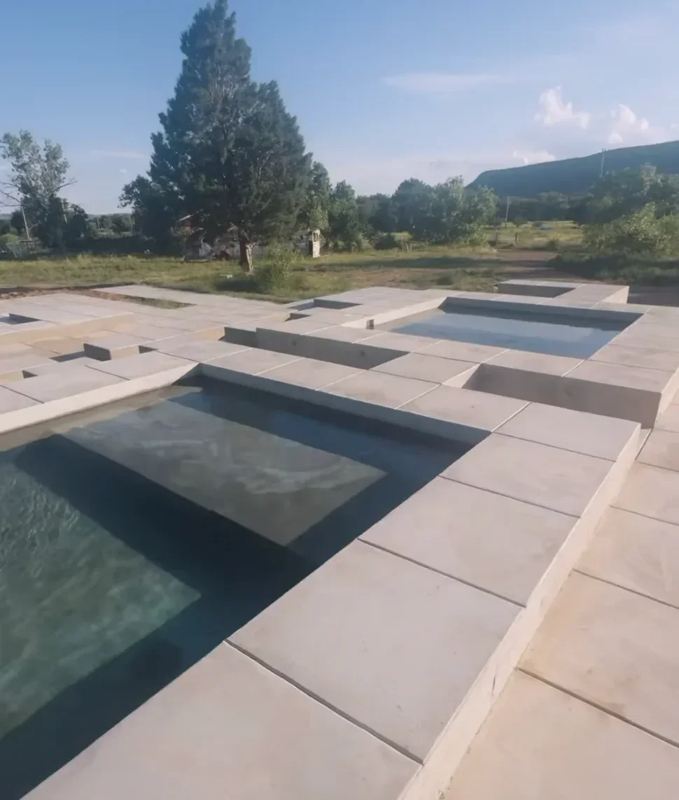Multiple rectangular concrete pools with water, surrounded by a paved area, with a backdrop of a large pine tree and distant hills under a blue sky.