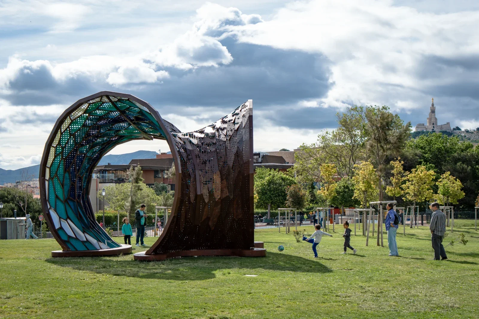 A park with a large metallic sculpture resembling a wave on the left, trees, people playing and walking, a grassy field, and a hill with a building on top in the background under a partly cloudy sky.