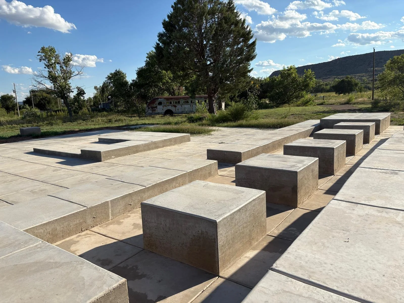 Concrete steps and platforms in an outdoor setting with grass, trees, a vintage bus, and mountains in the background under a partly cloudy sky.