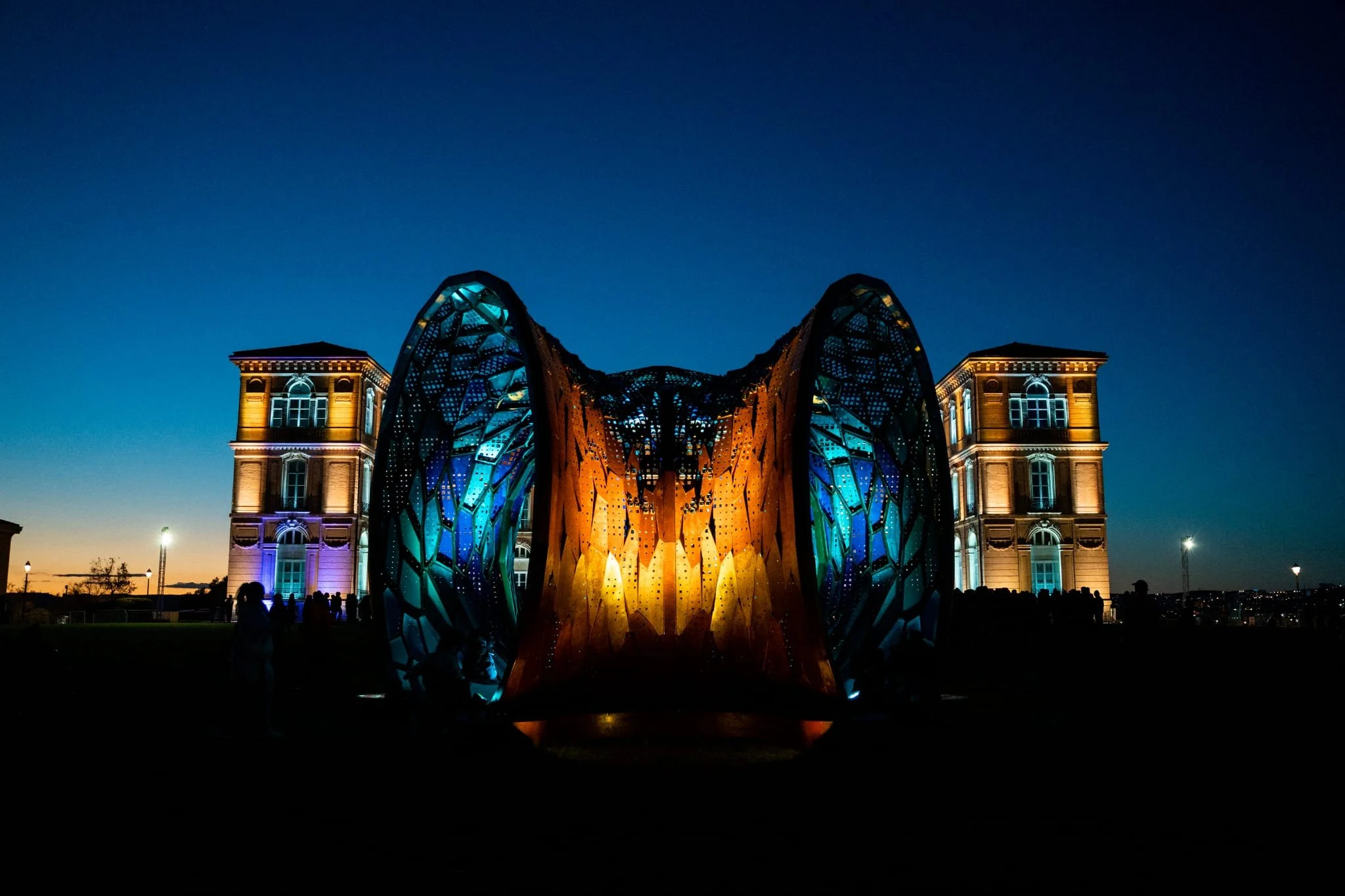 Nighttime view of a modern, illuminated sculpture resembling a butterfly or wings in front of a historic building with lit windows.