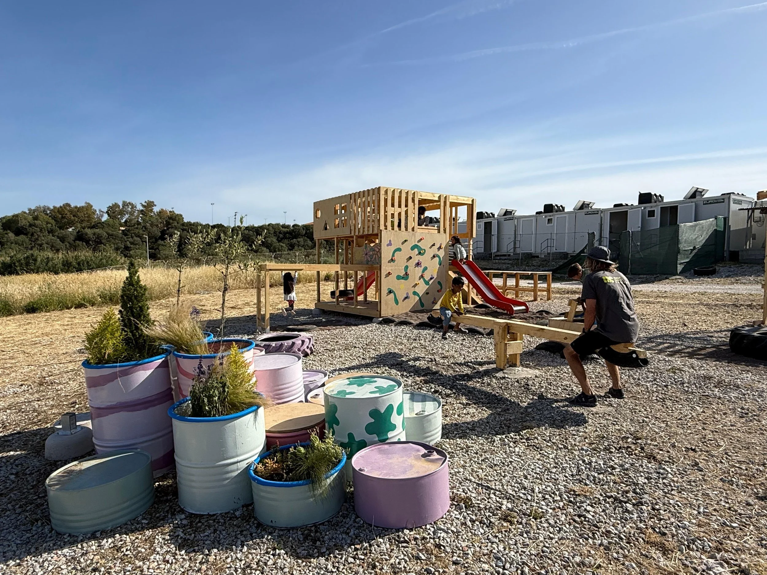 Children playing on a wooden climbing structure with a slide at an outdoor playground, surrounded by potted plants and gravel ground, with a backdrop of trees, hills, and a blue sky.
