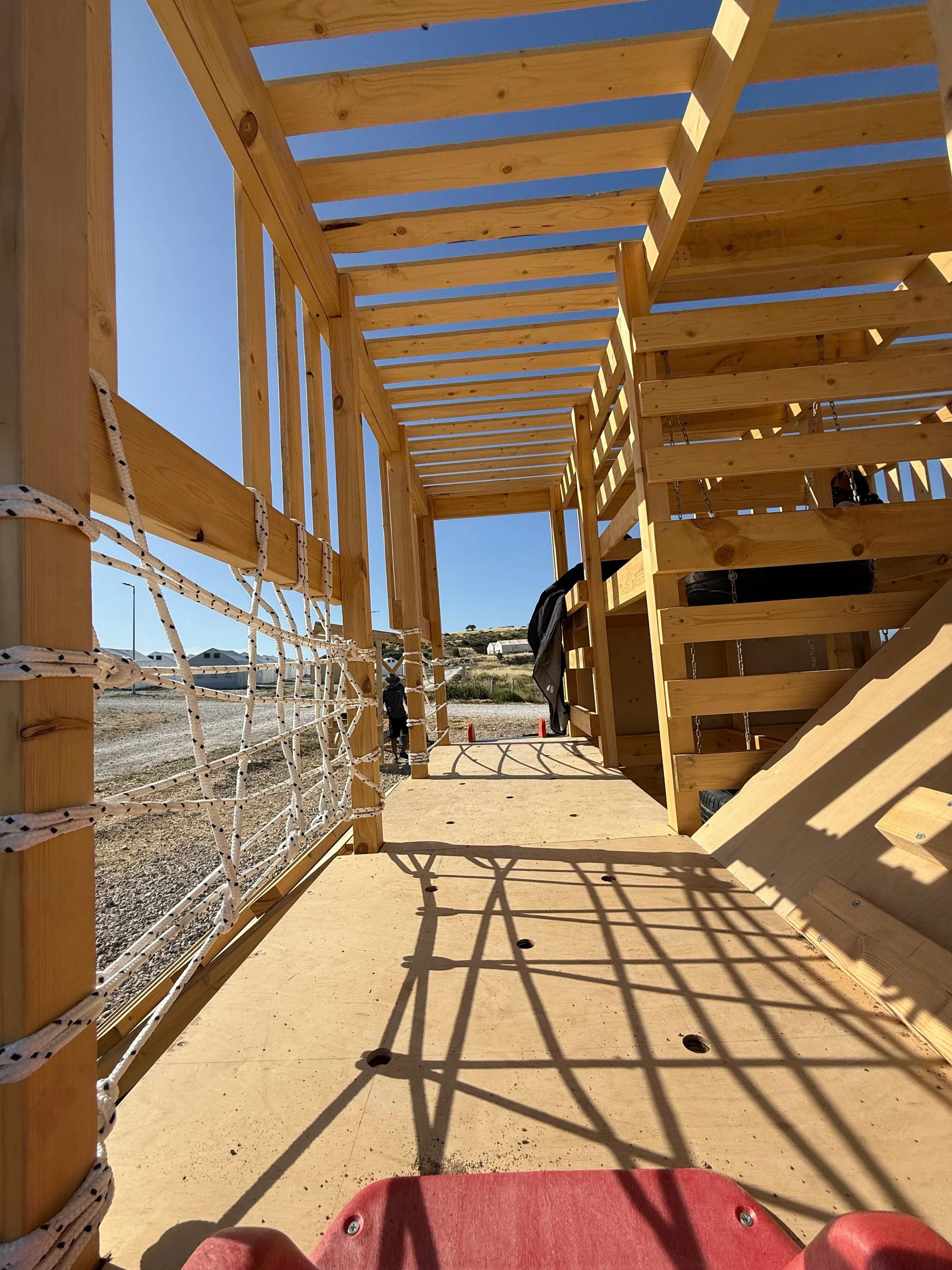 View from a child's perspective on a wooden play structure with ropes on the left and tires on the right. The structure is outdoors under a blue sky. In the background, there are some houses and a hill.