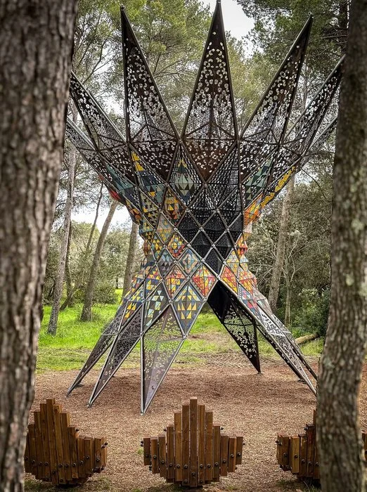 Large star-shaped metal sculpture with colorful geometric patterns, situated outdoors among trees, with wooden benches in the foreground.