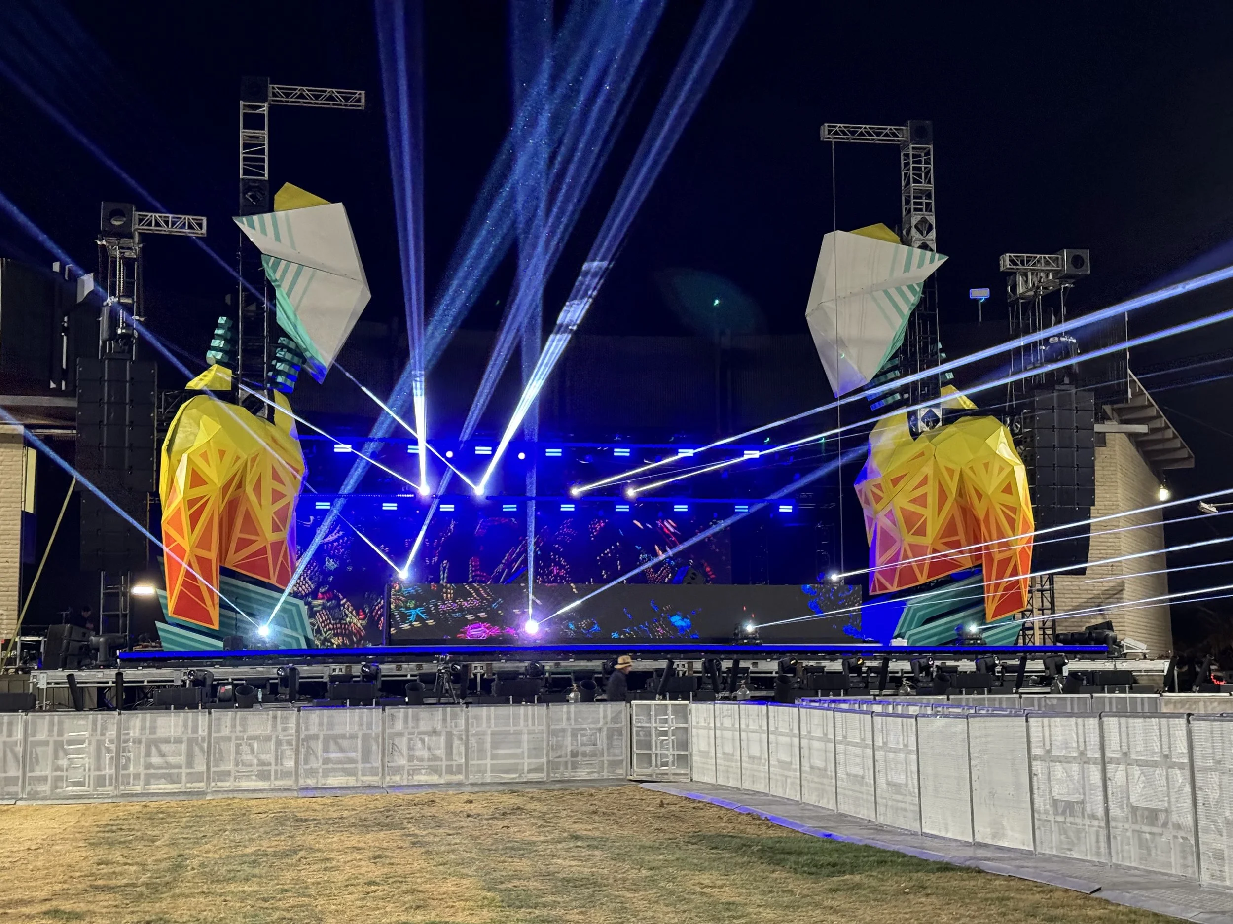 A concert stage at night with colorful geometric decorations and multiple blue and white laser light beams projecting into the sky. The stage is set with large speakers and lighting equipment, while a person stands in front on the ground.