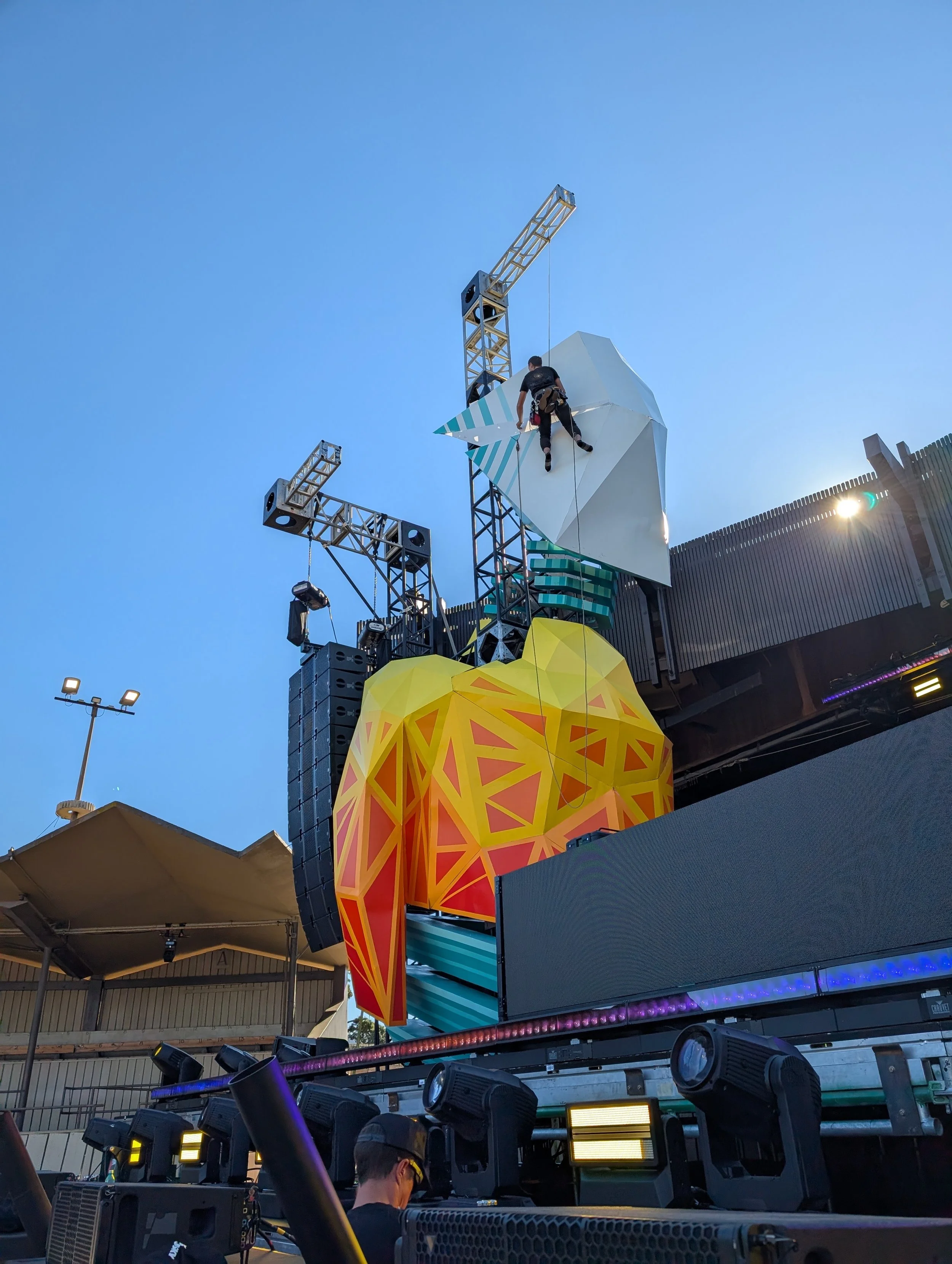 A person climbing on a green and yellow geometric sculpture, set against a blue sky with stage lighting and scaffolding in the background.