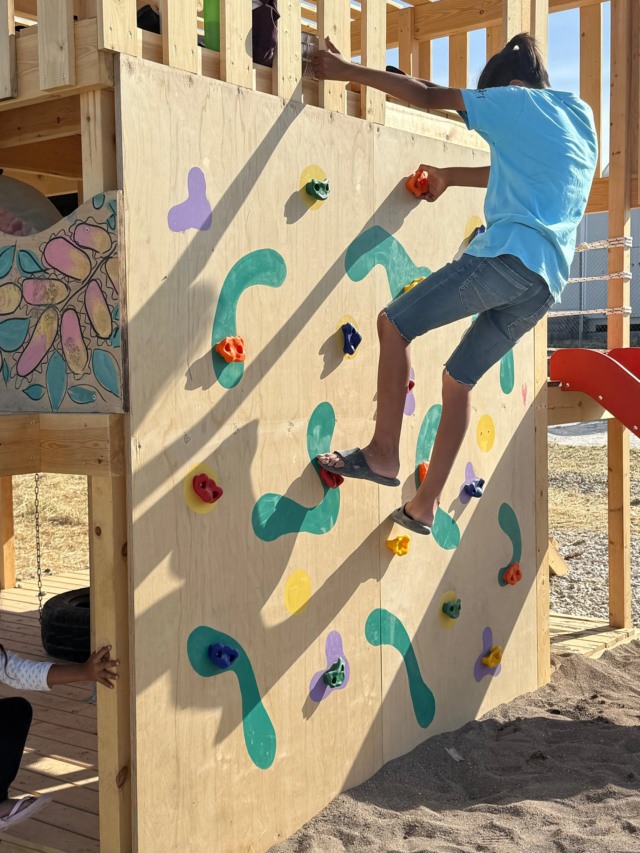 Child climbing on wooden outdoor climbing wall with colorful handholds at a playground.