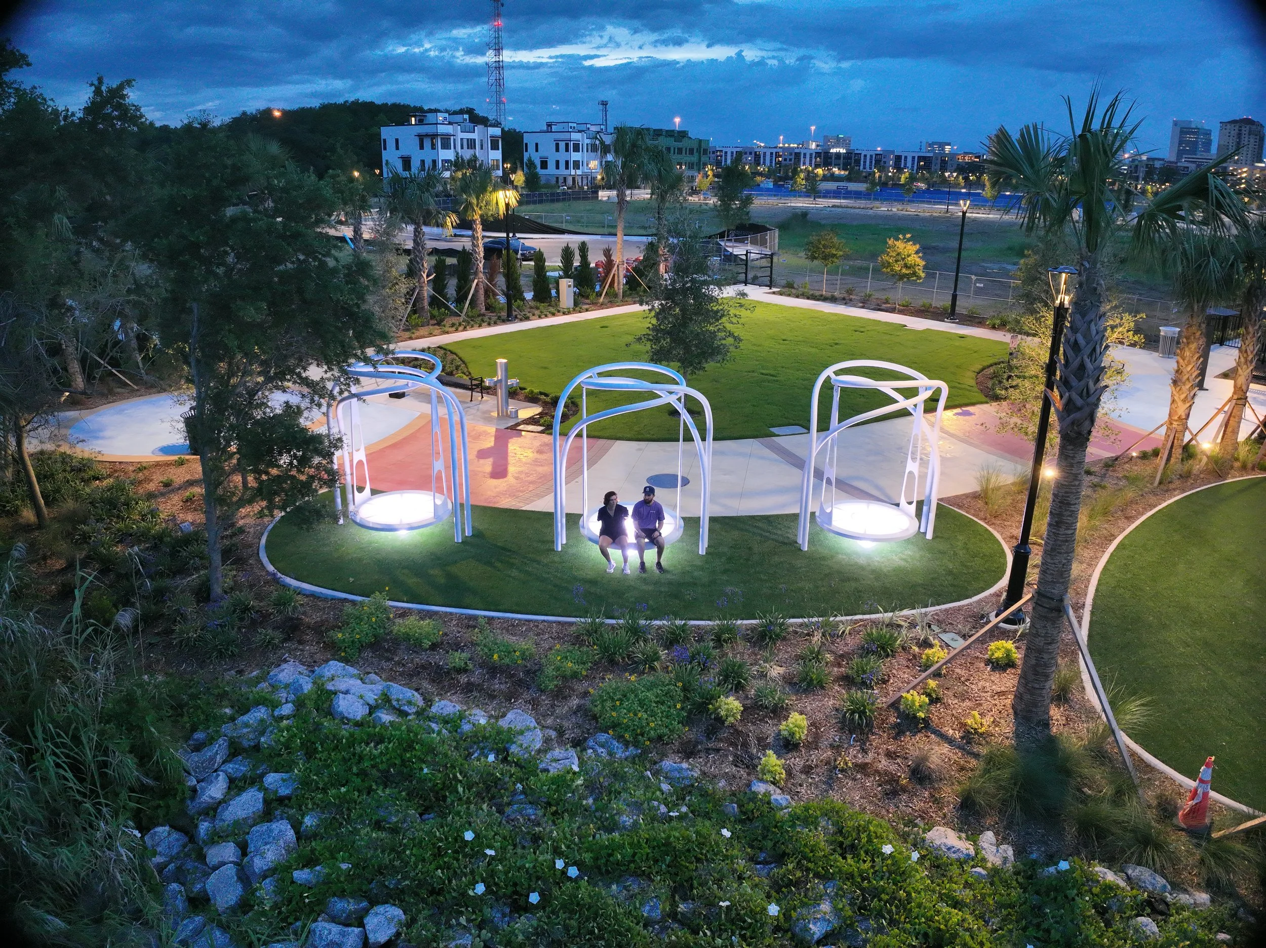 A park with a small grassy area, surrounded by trees and bushes, during twilight. There are modern residential buildings in the background, and a few streetlights illuminate the path and playground area. Two people are sitting on a swing in the foreground.