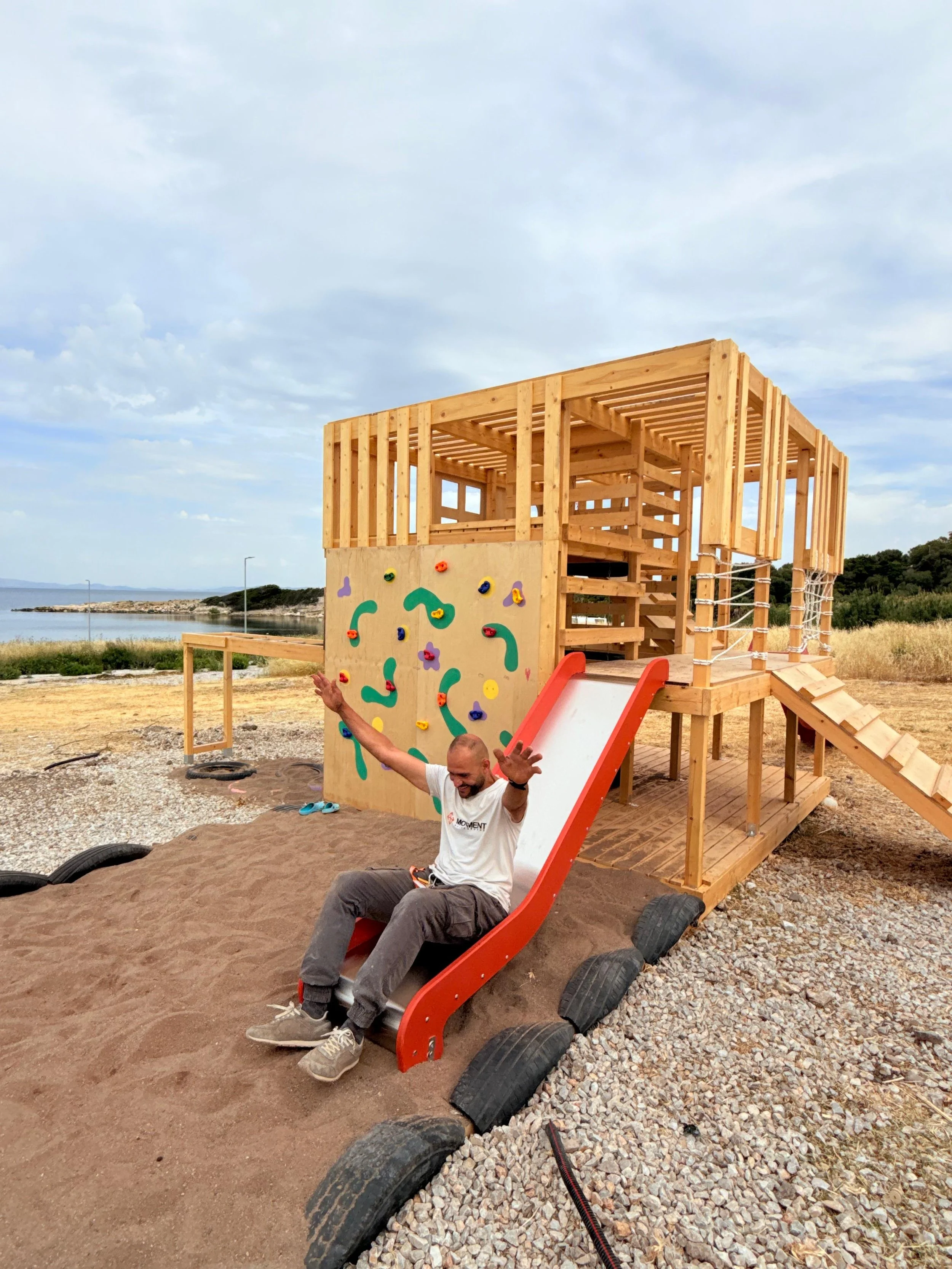 A man sitting on a slide with a construction playset of a wooden house and climbing wall behind him, outdoors near water and grassy area, with cloudy sky overhead.