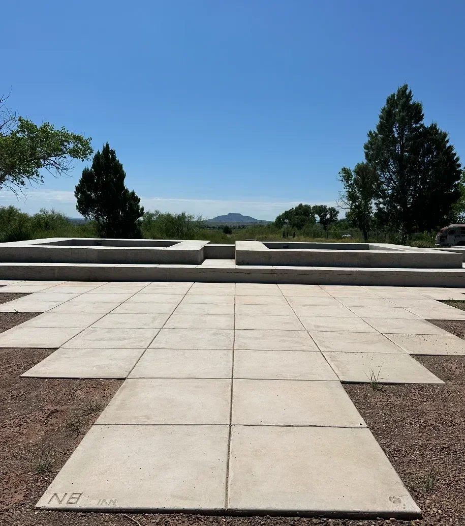 Concrete walkway leading to a memorial or monument with a flat top, situated outdoors with trees and a mountain in the background under a clear blue sky.