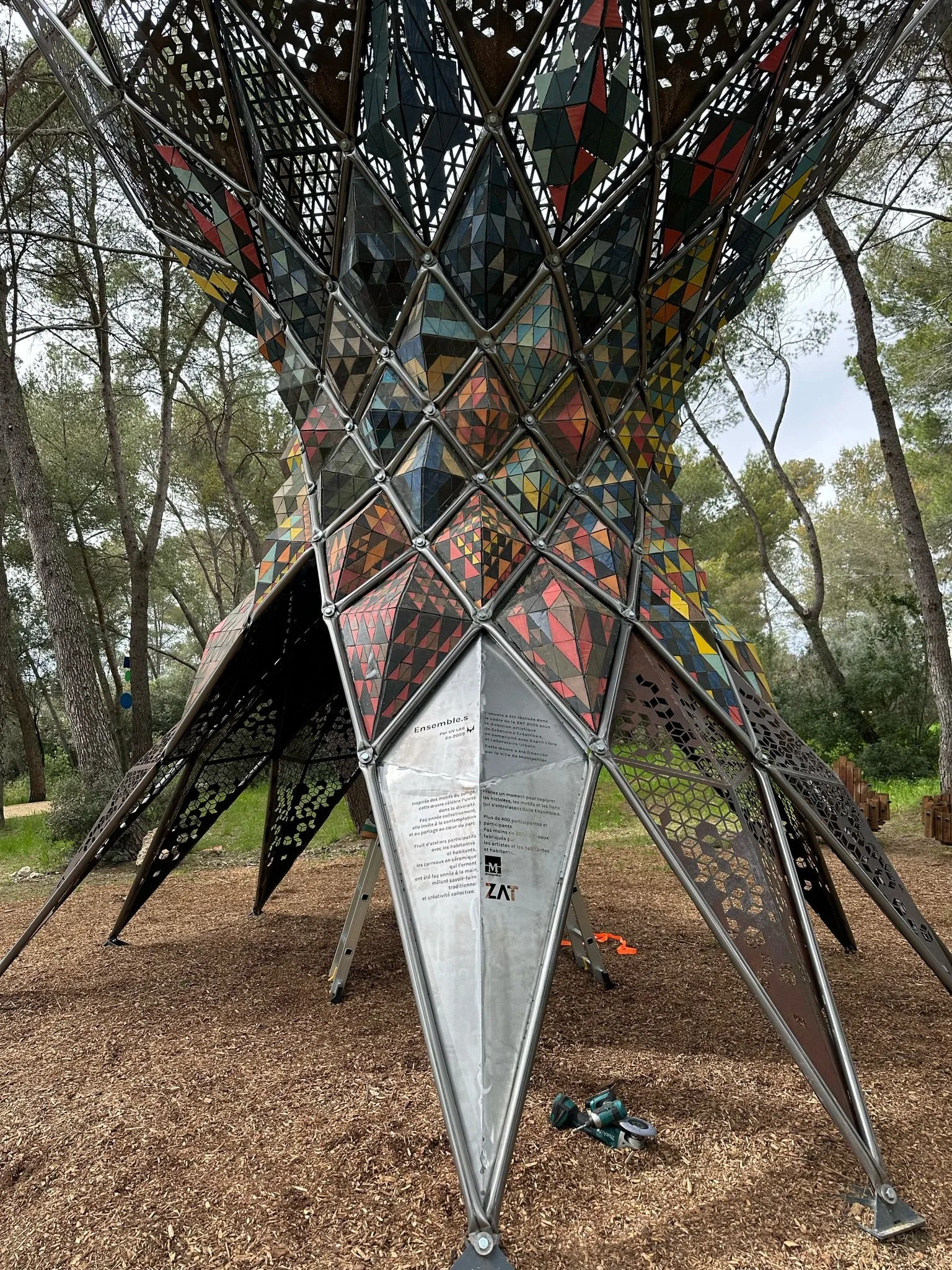 A colorful, geometric metal sculpture or installation in a park, surrounded by trees and dirt ground.
