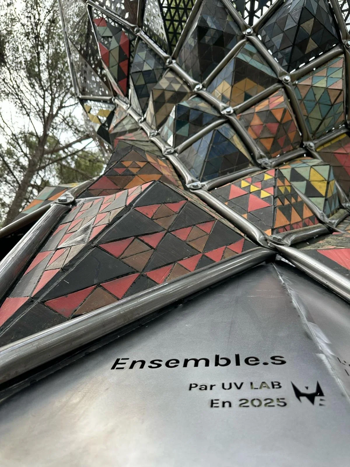 Close-up of a colorful mosaic sculpture with geometric patterns made of small tiles in various colors, metal framing, and a tree in the background.
