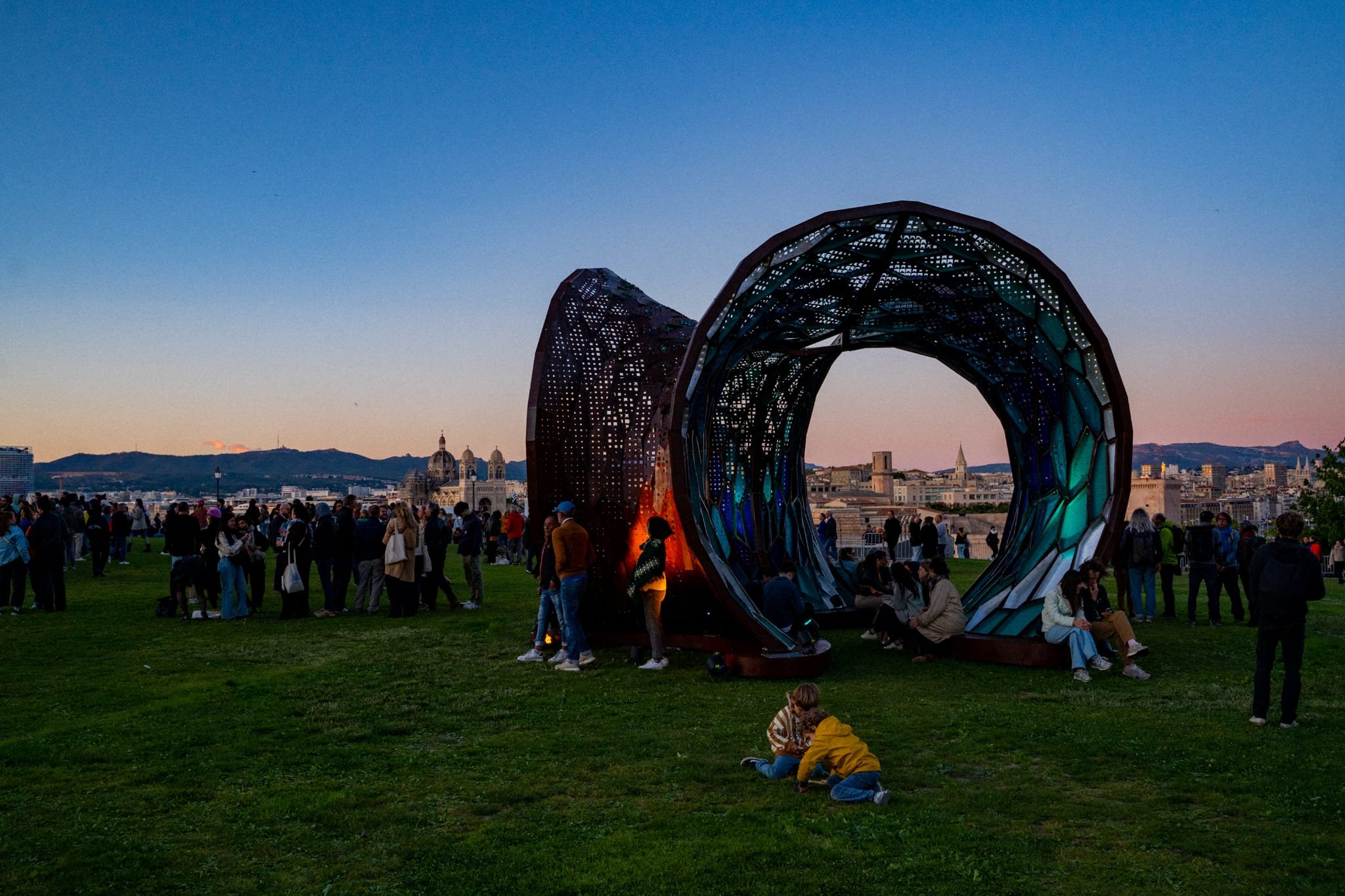 A large artistic sculpture resembling an eye with a detailed, colorful interior, set against a city skyline at dusk with a crowd of people gathered around in a park.