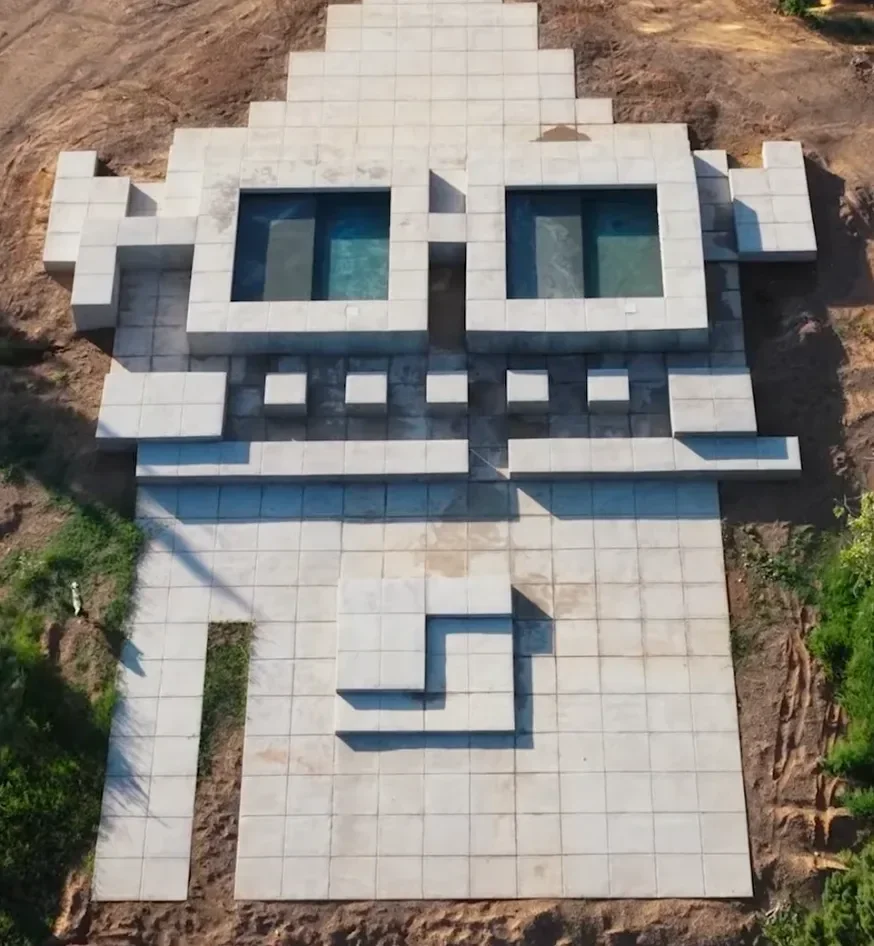 A large outdoor tiled area with a pixel art design of a character's face, featuring square eyes and a tongue, laid out on a dirt surface surrounded by greenery.